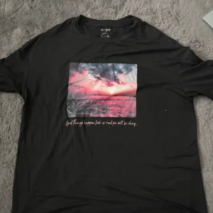 Svart t-shirt med tryck - Snygg svart t-shirt med ett färgglatt tryck av en solnedgång och texten 'Good things happen. Love is real. We will be okay.' Perfekt för en avslappnad stil.