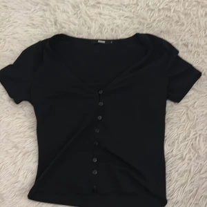 Svart kortärmad topp  - Snygg svart topp med korta ärmar och knappar framtill. Perfekt för en stilren look. Toppen har en v-ringad design som ger en elegant touch.