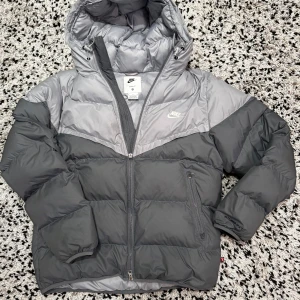 Grå dunjacka från Nike - Säljer en snygg grå dunjacka från Nike med huva och dragkedja. Jackan har en stilren design med två nyanser av grått och Nike-loggan på bröstet. Perfekt för kyliga dagar.