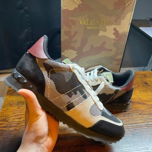 Valentino Rockrunners - Exklusiva Valentino Garavani Rockrunner sneakers i ett snyggt camouflage-mönster! En perfekt kombination av lyx och streetwear, med premiumkvalitet och de ikoniska nitarna vid hälen. |  Färg: Camouflage (svart, röd, vit) 🖤🤍❤️ | Material: Mix av mocka, mesh och läder 🏆| Skick: Nästan nya🙌 | Storlek: 44 👌| Box: Originalbox medföljer 📦✨ | 🚚 Leverans: Skickas snabbt via PostNord eller instabox | 💬 Skicka ett meddelande vid frågor!