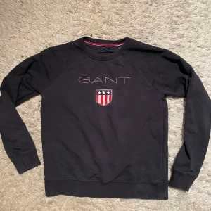 Mörkblå gant sweatshirt  - skick 9/10 .Storlek :176 passar ungefär XS.    Säljer en stilren svart sweatshirt från GANT med broderad logotyp och emblem på bröstet. Tröjan har långa ärmar och en rund halsringning med röd och vit detalj på insidan. Perfekt för en avslappnad look. Hör av er vid fler funderingar!