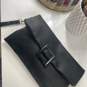 Svart kuvertväska med spänne - Snygg svart kuvertväska i syntetmaterial med ett stilrent fyrkantigt spänne på framsidan. Väskan har en praktisk magnetknapp för enkel öppning och stängning. Perfekt för att bära med sig det nödvändigaste.