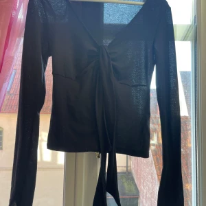 Svart långärmad topp från Gina Tricot - Snygg svart långärmad topp från Gina Tricot med vida armar, V ringning och knytning framtill. Funkar som basic och även festlig. Tillverkad i ett lätt och transparent material som ger en elegant touch. Man kan knyta där framme.