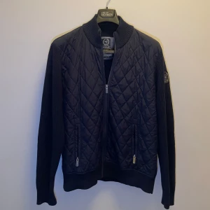 Mörkblå quiltad jacka från State of Art - Snygg mörkblå quiltad jacka från State of Art med ribbad krage och ärmar. Jackan har en dragkedja framtill och ett märke på ärmen. Perfekt för kyligare dagar.