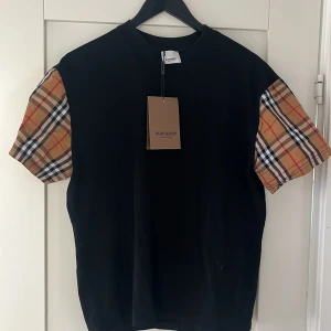 Burrberry T-shirt  - Snygg svart t-shirt från Burberry med deras ikoniska rutmönster på ärmarna. Perfekt för en stilren look med en touch av klassiskt mönster. T-shirten har en normal passform och är kortärmad. Är L men passar M