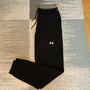 Under Armour träningsbyxor - Snygga svarta träningsbyxor från Under Armour med elastisk midja och dragsko. Byxorna har en diskret logga på benet och en praktisk ficka med dragkedja baktill. Perfekta för träning och avslappnade dagar.