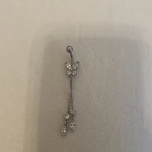 Silverfärgad navelpiercing med fjäril och kedjor - Elegant navelpiercing i silverfärg med en glittrig fjäril och hängande kedjor med små kristaller. Perfekt för att ge en extra touch till din stil.