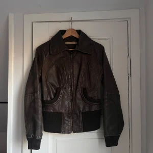 Brun skinnjacka från Vero Moda - Snygg brun skinnjacka från Vero Moda med dragkedja framtill och ribbade muddar vid ärmslut och nederkant. Jackan har en klassisk krage och två fickor framtill. Perfekt för en cool och stilren look.
