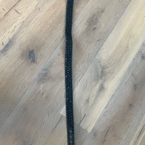 Svart glittrigt bälte med nitar - Snyggt svart bälte med glittriga detaljer och nitar. Bältet har en stor spänne dekorerad med svarta stenar och en cool design. Perfekt för att ge din outfit en extra edge.
