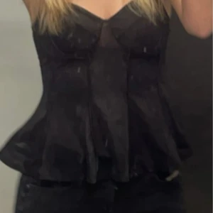 Svart korsett med volangdetaljer - Snygg svart korsett med volangdetaljer och en elegant passform. Perfekt för en kväll ute eller en speciell tillställning. Den har en v-ringad design och är tillverkad i ett glansigt material som ger en lyxig känsla.