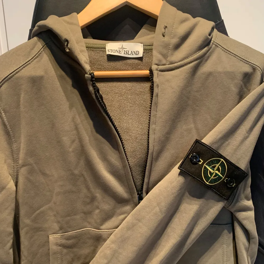 Säljer en snygg gr hoodie från Stone Island. Den har en dragkedja framtill och en klassisk huva. På ärmen finns det ikoniska Stone Island-märket. Perfekt för höst och vår, och den är i bra skick. Passar både till vardags och lite mer avslappnade till. Tröjor & Koftor.