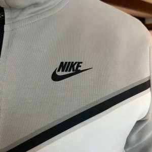 Grå och vit hoodie från Nike - Säljer en snygg och bekväm grå och vit hoodie från Nike. Den har en dragkedja framtill och en huva med dragsko. Perfekt för både träning och vardagsbruk. Den har en normal passform och är långärmad med en cool svart detalj på ärmen.