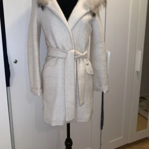 Vit kappa med pälskrage - Säljer en superfin vit kappa med pälskrage och bälte i midjan. Den är perfekt för vintern och hösten, och har en mysig och elegant stil. Kappan är långärmad och har en huva med äkta päls  som ger en lyxig känsla. 