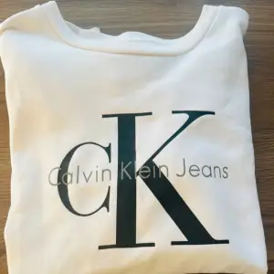 Säljer en snygg vit sweatshirt från Calvin Klein Jeans. Den har en stor CK-logga på framsidan och är supermjuk och bekväm. Perfekt för en avslappnad stil eller att mysa i hemma. Passar till alla tillfällen!