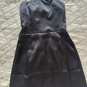 Svart axelbandslös klänning - Säljer en elegant svart axelbandslös klänning i satin. Perfekt för festliga tillfällen! Klänningen har en smickrande passform och en elastisk rygg för extra komfort. Den är i nyskick och redo att bäras till nästa stora event.