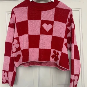 Röd och rosa stickad tröja med hjärtmönster - Fin stickad tröja i rött och rosa med ett schackrutigt mönster med hjärtan och blommor. Använd ett fåtal gånger men har inga defekter. Storlek S från Only.