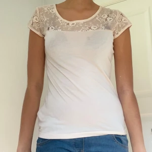 Vit topp med spetsdetaljer - Säljer en superfin vit topp i storlek 38. Den har korta ärmar och en vacker spetsdetalj över axlarna och övre delen av ryggen. Toppen är perfekt för sommardagar och har en normal passform. Den knäpps med små knappar i ryggen för en extra söt touch.