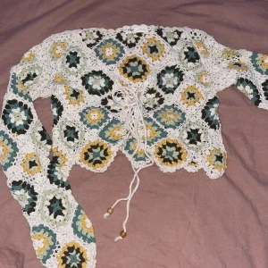 Flerfärgad virkad topp - Säljer en superfin virkad topp med ett unikt mönster i grönt, gult, vitt och svart. Toppen är croppad och har långa ärmar med snörning framtill. Perfekt för vår och sommar, och den ger en härlig boho-vibe. Passar perfekt till festivaler eller en dag på stan!