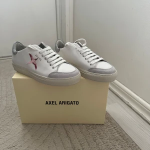 Vita sneakers från Axel Arigato - Säljer ett par snygga vita sneakers från Axel Arigato. De har en cool broderad fågel i rosa på sidan och grå detaljer vid tån och hälen. Skorna är i nyskick och kommer med originalförpackning och dustbag. Perfekta för både vardag och fest!
