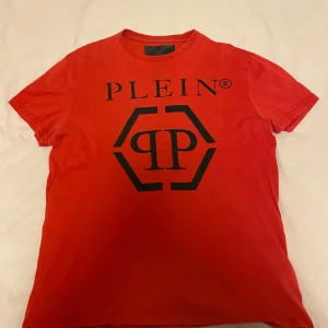 Philipp plein - Säljer en snygg röd t-shirt från Plein med ett stort svart tryck på framsidan. Den är kortärmad och perfekt för både vardag och fest. T-shirten är i bra skick och har en normal passform. Perfekt för dig som vill ha något stilrent men ändå med en touch av lyx.