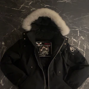 Svart vinterjacka från Moose Knuckles - Säljer en svart vinterjacka från Moose Knuckles i nyskick. Jackan har en fluffig pälsfodrad huva och dragkedja framtill. Perfekt för kalla vinterdagar. Kommer med originalförpackning och NFC-tag för äkthetsbevis.