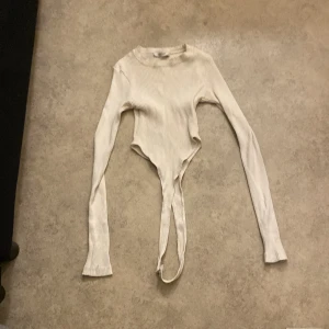 Vit ribbad topp från QU STYLE - Säljer en vit ribbad topp från QU STYLE i storlek S. Toppen har långa ärmar och en unik design med knytband i midjan. Perfekt för både vardag och festliga tillfällen. Den är i mycket bra skick och redo för en ny ägare!