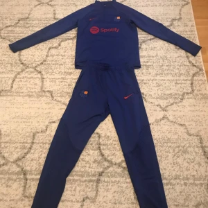 Blå träningsset från Nike - Säljer ett blått träningsset från Nike med långärmad tröja och byxor. Tröjan har en dragkedja vid halsen och tryck med Spotify-logga. Perfekt för fotbollsträning eller annan sportaktivitet. Har använts ett par gånger och har inga skador. Det originala pricet är 800kr men säljer för 300kr.