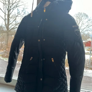 Svart quiltad jacka från Rockandblue - Säljer en svart quiltad jacka från Rockandblue i mycket bra skick. Den har en huva och dragkedja framtill, samt en snygg detalj med en ficka och logga på ärmen. Perfekt för kyliga höst- och vinterdagar. Storleken är dam xs