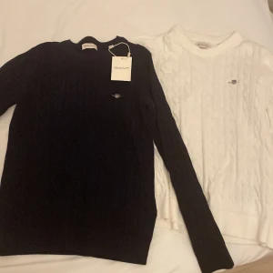 Svart och vit kabelstickad tröja från GANT - Säljer två snygga kabelstickade tröjor från GANT i storlek 146-152. En är klassiskt svart och den andra är stilrent vit. Båda har GANT-loggan broderad på bröstet och är i nytt skick med etikett kvar. Perfekta för höst och vinter!
