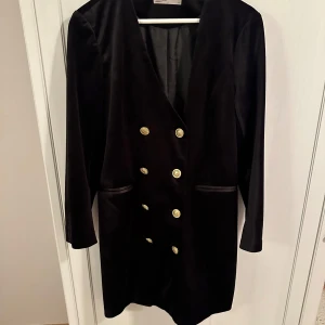 Svart klänning från Vero Moda - Snygg svart klänning från Vero Moda i storlek 40. Den har en elegant dubbelknäppt design med guldknappar och långa ärmar. Perfekt för höst och vinter, och passar bra till festliga tillfällen. Materialet är i sammet. Perfekt till jul och nyår. 