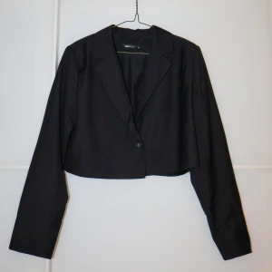 Svart croppad kavaj från Gina Tricot - Säljer en stilren svart croppad kavaj från Gina Tricot i storlek 38. Perfekt för både vardag och fest! Kavajen har en enkel knapp framtill och långa ärmar. Den är i mycket bra skick och passar perfekt till höst- och våroutfits.