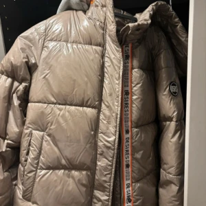 Beige pufferjacka från Desires - Säljer en snygg beige pufferjacka från Desires. Jackan har en glansig finish och en dragkedja framtill. Den är perfekt för kyliga dagar och har en logga på ärmen. Superbekväm och stilren för både vardag och fest! Jackan är lite stor i storleken, sitter som en S