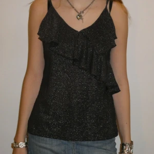 Svart glittrig festtopp med volangdetalj - Snygg, svart, glittrig topp med volangdetalj framtill. Perfekt på fest eller liknande. Storlek M. 