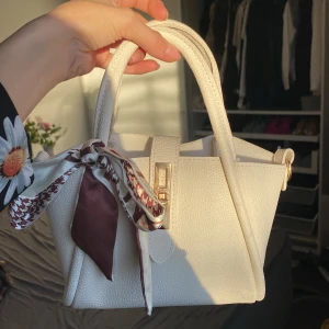 Vit handväska med scarfdetalj - Säljer en superfin vit handväska i syntetmaterial med en snygg scarfdetalj knuten runt handtaget. Väskan har en elegant design med guldfärgade detaljer och en avtagbar axelrem. Perfekt för både vardag och fest!