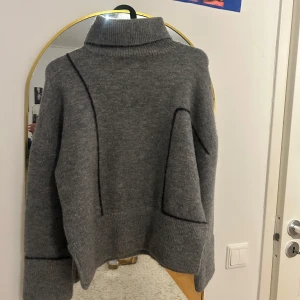 Grå stickad polotröja - Säljer en supermysig grå stickad polotröja med en unik design. Den har en hög krage och långa ärmar, perfekt för kyliga dagar. Tröjan har en cool asymmetrisk detalj framtill som ger den en modern touch. Perfekt för både höst och vinter!