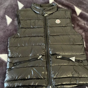 Svart dunväst från Moncler - Säljer en snygg svart dunväst från Moncler i mycket bra skick. Västen har en glansig yta med dragkedja och två praktiska fickor framtill. Perfekt för höst och vår när du vill hålla dig varm och stilren. Passar bra till både vardag och fest!