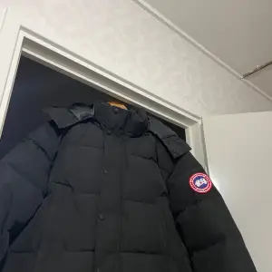 Säljer en svart dunjacka från Canada Goose i storlek M. Jackan är perfekt för kalla vinterdagar och har en klassisk design med huva och knappar. Den är i mycket bra skick och håller dig varm och bekväm. Passar både till vardags och mer formella tillfällen.