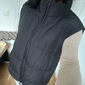 Svart väst - Säljer en svart dunväst från Monki i mycket bra skick. Västen har en dragkedja framtill och är perfekt för höst och vinter. Den är både stilren och varm, vilket gör den idealisk för kyligare dagar. Oversized passform. Säljer pga den ej kommer till användning :)