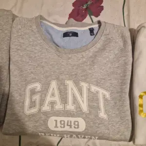 Säljer en snygg grå sweatshirt från GANT i storlek XL. Den har ett klassiskt tryck med 'GANT 1949' på framsidan. Perfekt för en avslappnad stil och passar bra till både jeans och chinos. Tröjan är i mycket bra skick och redo för nya äventyr!