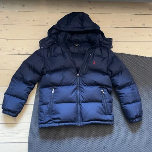 Mörkblå dunjacka från Polo Ralph Lauren - Säljer en snygg mörkblå dunjacka från Polo Ralph Lauren storlek Barn L men passar XXS runt 165-175. Jackan har en praktisk huva och dragkedja framtill. Den är perfekt för kalla dagar och har en klassisk design med det ikoniska röda Polo-logotypen på bröstet. Perfekt för vintern! ❄️