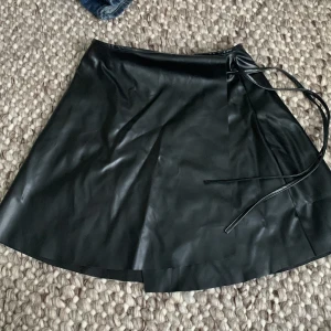 Svart omlottkjol i skinnimitation - Snygg svart omlottkjol i skinnimitation som ger en cool look. Kjolen har en knytning i sidan för en justerbar passform. Perfekt för både vardag och fest! 🖤