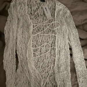 Vit spetskofta från Gina Tricot - Säljer en superfin vit spetskofta från Gina. Den är perfekt för våren och sommaren med sitt lätta och luftiga material. Koftan har långa ärmar och en snygg snörning framtill som ger en extra detalj. Perfekt att ha över en topp eller klänning för en chic look!