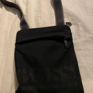 Svart axelväska från Boss - Säljer en stilren svart axelväska från Boss. Väskan har en praktisk dragkedja och justerbar axelrem. Perfekt för vardagsbruk eller när du är på språng. Den är i bra skick och redo för en ny ägare!