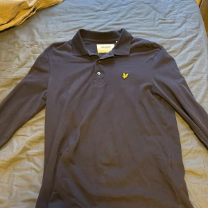 Mörkblå pikétröja från Lyle & Scott - Säljer en stilren mörkblå pikétröja från Lyle & Scott i ekologiskt material. Tröjan har långa ärmar och en klassisk krage med knappar. Perfekt för både vardag och lite finare tillfällen. Den har en liten gul logga på bröstet som ger en snygg detalj. Passar perfekt till höst och vår!
