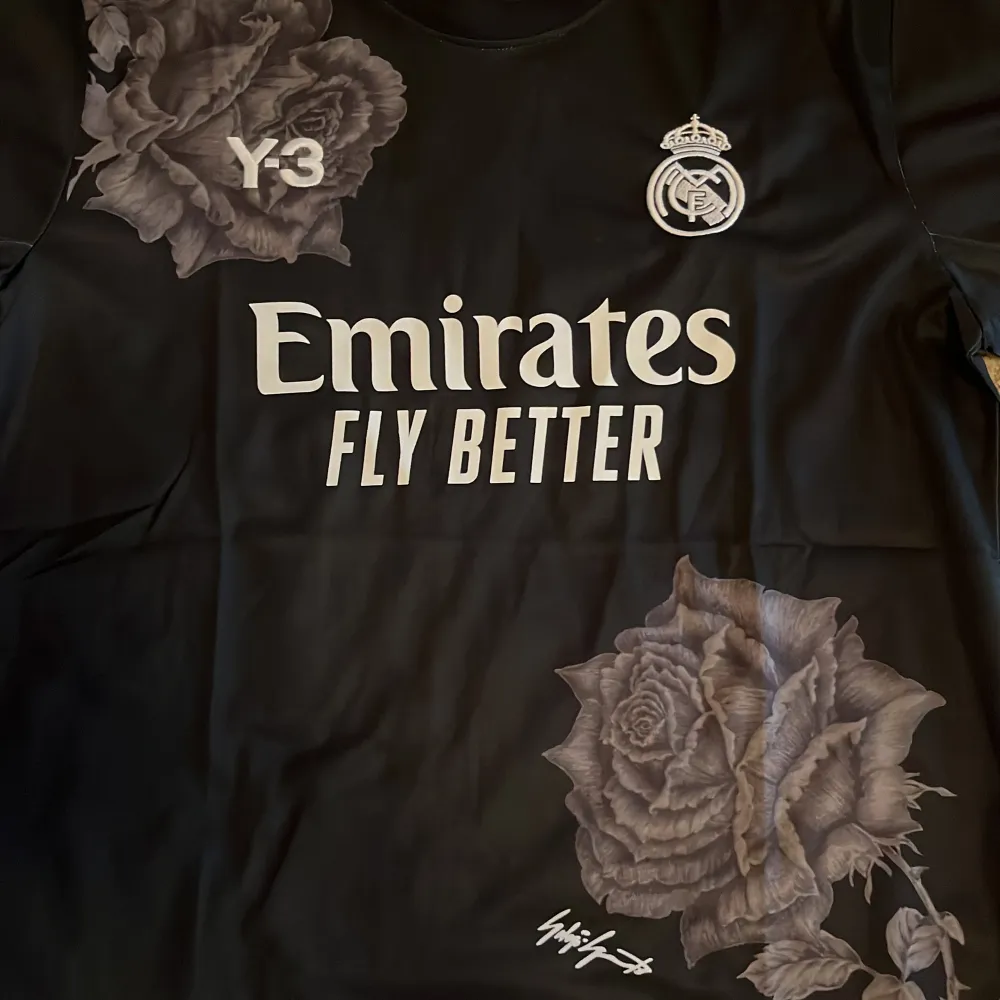 Säljer en svart Real Madrid fotbollströja i samarbete med Y-3. Tröjan har ett unikt blommönster och texten 'Emirates Fly Better' på framsidan. Den är i nytt skick och har korta ärmar. Perfekt för samlare eller fans av laget! 🌹⚽. T-shirts.