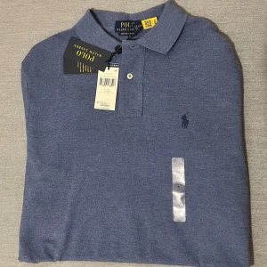 Blå pikétröja från Ralph Lauren - Säljer en snygg blå pikétröja från Ralph Lauren i nytt skick. Tröjan har en klassisk krage och knappar framtill. Perfekt för både vardag och lite finare tillfällen. Den är i en skön bomullskvalitet och har den ikoniska loggan broderad på bröstet.