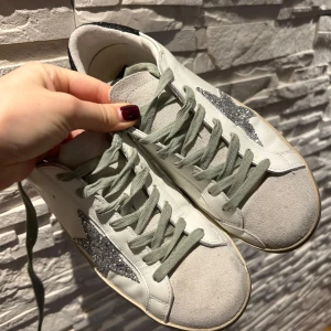 Golden Goose sneakers med glittrig stjärna - Säljer ett par snygga Golden Goose sneakers i vitt skinn med en glittrig silverstjärna på sidan. Skorna har grå snörning och svart glittrig häl. Perfekta för att ge din outfit lite extra bling! 