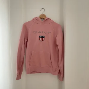 Rosa hoodie från GANT - Säljer en superfin rosa hoodie från GANT i bra skick. Den har en stor ficka fram och ett snyggt tryck med GANT-loggan och ett emblem. Perfekt för höst och vår när du vill vara både bekväm och stilren. Passar till allt från jeans till mysbyxor!