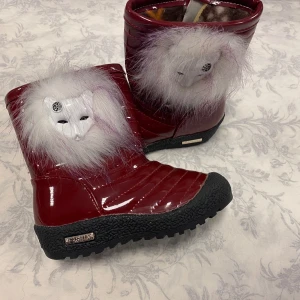 Vinröda vinterstövlar med pälsdetalj - Säljer ett par unika vinröda vinterstövlar med vit pälsdetalj och maskmotiv. De har en robust svart sula och är fodrade med mönstrat tyg för extra värme. Perfekta för kalla vinterdagar och ger en cool look med sin glansiga yta.                                                           Stlk kvar 27,28,30   Med fleece inuti,, rekommenderar att välja en Stlk större 