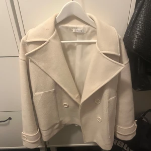 Beige kappa från Designs by Si - Säljer en superfin beige kappa från Designs by Si. Den har en klassisk look med stora slag och knappar framtill. Perfekt för höst och vår! Jackan är i mycket bra skick och har en normal passform. Den är långärmad och har praktiska fickor.